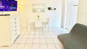 Uma sala de estar com televisão branca, mesa de jantar com cadeiras, sofá e um quadro na parede.
