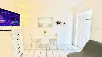 Uma sala de estar com televisão branca, mesa de jantar com cadeiras, sofá e um quadro na parede.