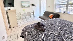 Quarto com cama, sofá, escrivaninha com cadeira, televisão e janela com persianas.