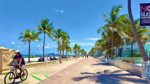 A paved walkway with palm trees and beach umbrellas, a cyclist, and a sign prohibiting glass containers on the beach. - Southwinds #4 for 2, Steps to Beach & Boardwalk (Hollywood)