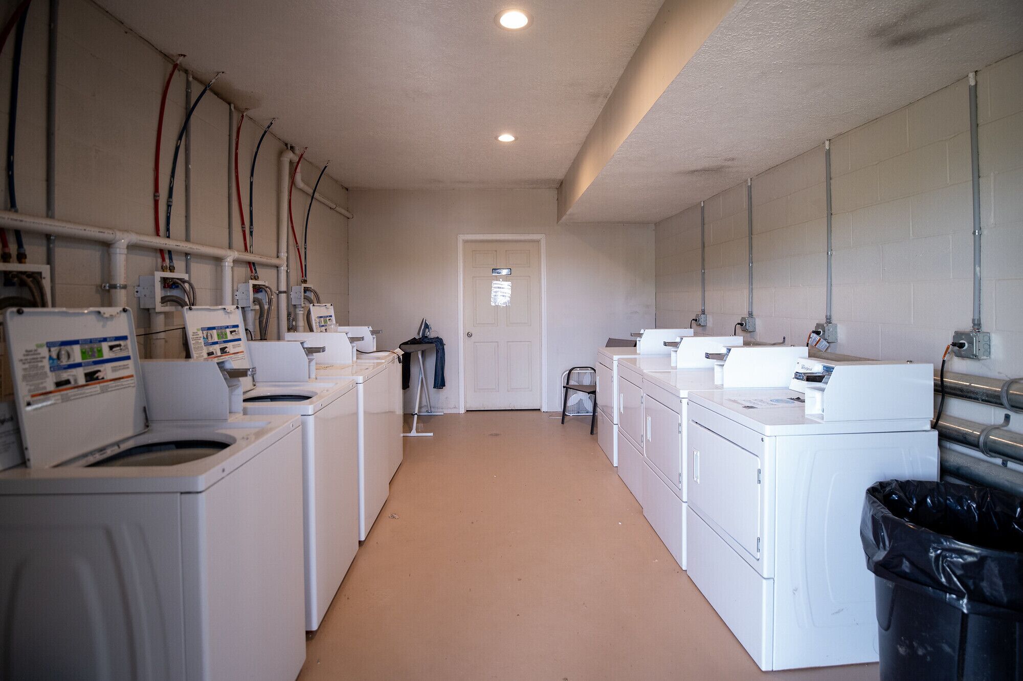A laundry facility with multiple washing machines and dryers, a door, and visible plumbing on the walls.