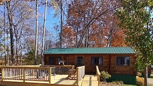 A wooden cabin with a green roof, surrounded by trees and a fenced deck area.