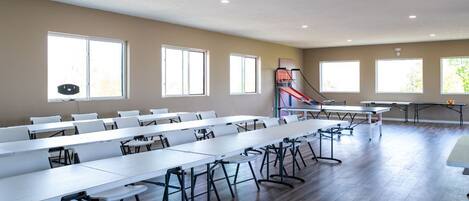 A spacious room with long tables and chairs, large windows, and a basketball hoop.