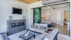 A modern living room with a fireplace, a television, and a large window overlooking a green space. - Costal Chic Vila w/Private Pool/Spa --5 min from the Beach! (Naples)