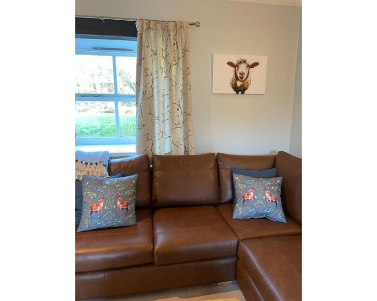 A brown leather sofa with two decorative pillows, a window with a view of greenery, and a framed picture of a sheep on the wall.