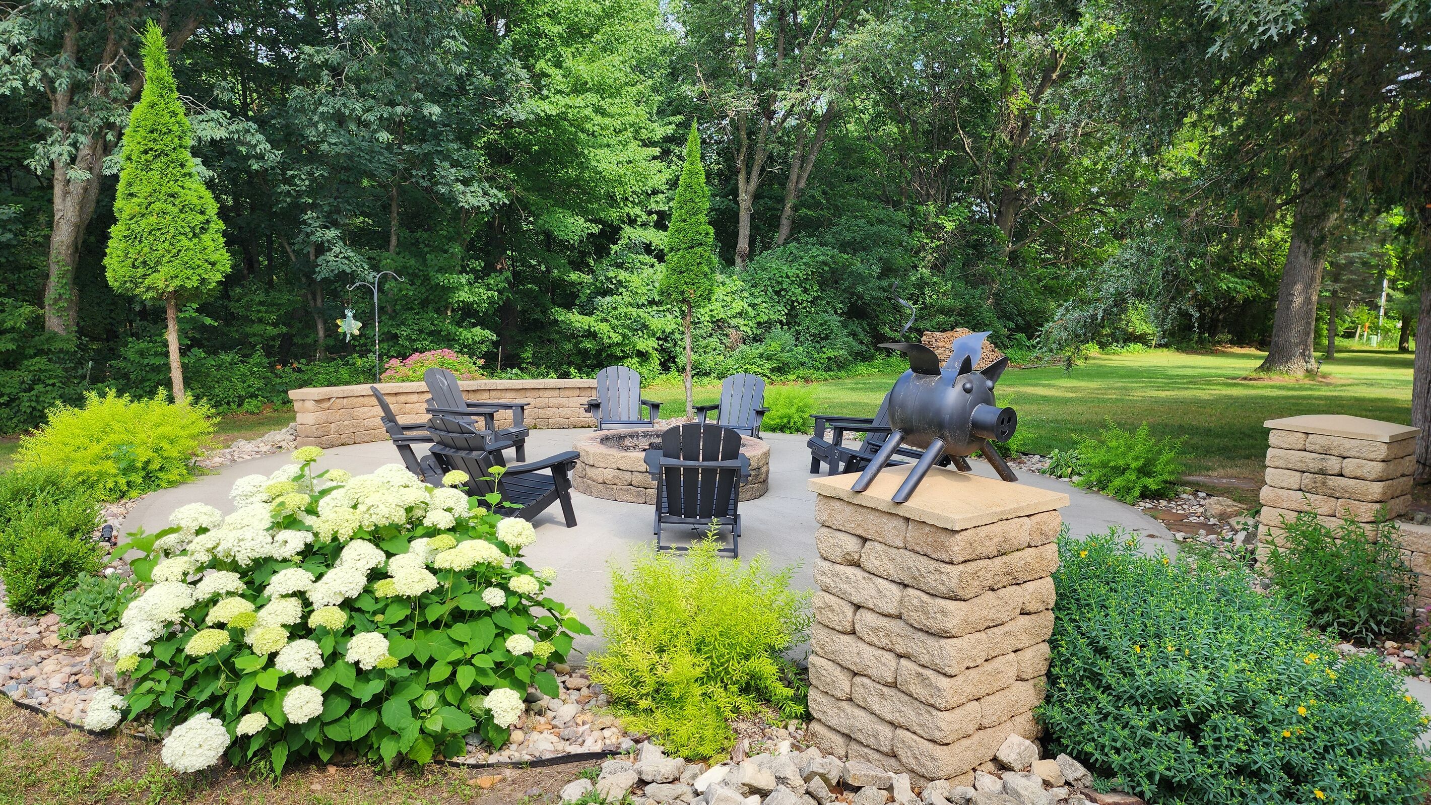 An outdoor seating area with Adirondack chairs, a stone fire pit, and a decorative pig sculpture.