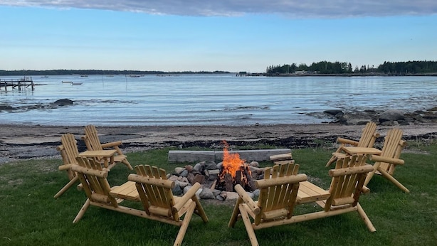 A lakeside setting with a fire pit and wooden chairs arranged around it. - Have an unforgettable getaway at a family sized beach house with private beach! (Steuben)