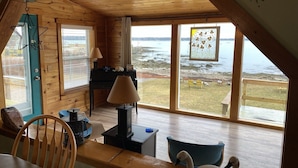 A cozy room with wooden walls and a sloped ceiling, featuring a table with chairs, a lamp, and a swan decoration. Large windows offer a view of a rocky shoreline and a body of water. - Have an unforgettable getaway at a family sized beach house with private beach! (Steuben)