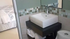 A bathroom with a white sink, a toilet, and a mirror. There is a bed with white bedding in the adjacent room. - Family Studio with all the comforts, centrally located. (Jalapa)