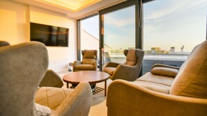 A modern living room with a round coffee table and beige sofas, large windows with a city view, and a flat-screen TV mounted on the wall.