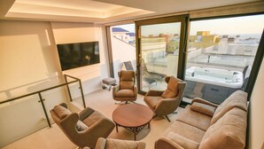 A modern living room with a sofa, armchairs, and a coffee table. There is a large window with a view of buildings and a balcony.