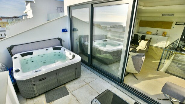 A modern balcony with a hot tub, a glass door leading to a kitchen, and a seating area with a chair and table.