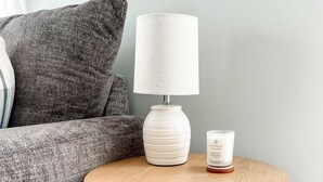 A round wooden table with a woven basket seat, a white lamp, and a candle on a grey textured sofa.