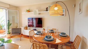 A dining area with a round wooden table set for four, surrounded by wicker chairs. A television is mounted on the wall above the table, and there are shelves with decorative items and a large woven light fixture above. - Balmins Surf by Hello Homes Sitges (Sitges)