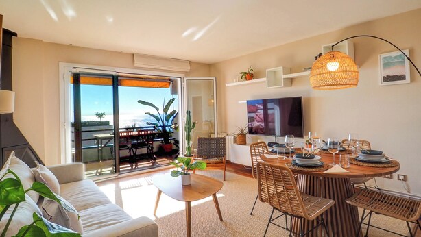 A modern living room with a sofa, dining table, chairs, and a TV. There is a balcony with a view of the sea. - Balmins Surf by Hello Homes Sitges (Sitges)