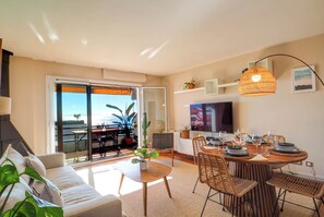 Un salon moderne comprenant un canapé, une table à manger, des chaises et un téléviseur. Il y a également un balcon avec vue sur la mer.