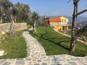 A stone pathway leading to a yellow house with a red roof, surrounded by trees and a stone wall.