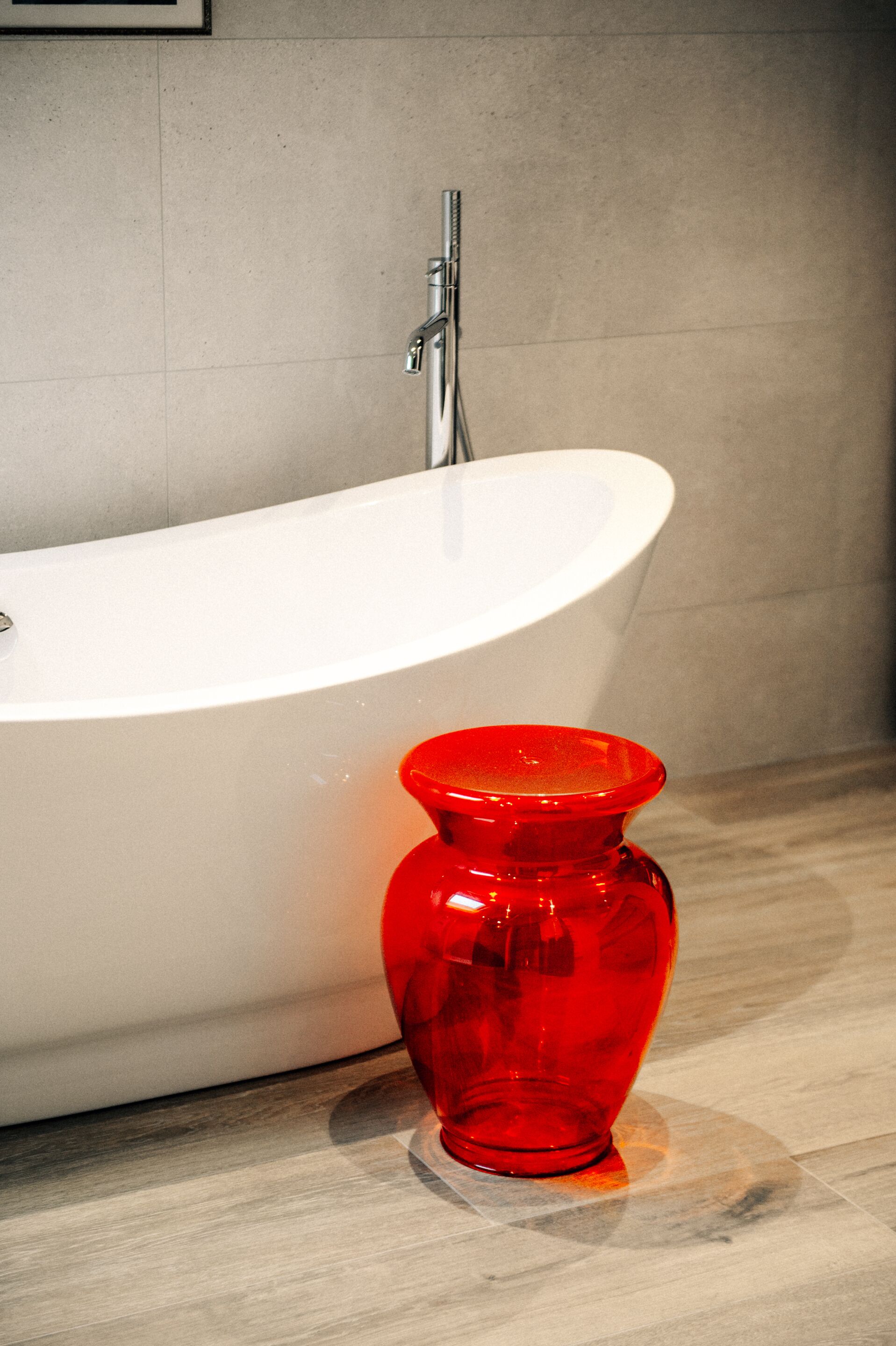 A white bathtub with a modern faucet, a red glass vase on a marble surface, and a tiled wall in the background.
