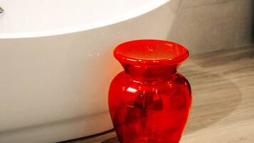 A white bathtub with a modern faucet, a red glass vase on a marble surface, and a tiled wall in the background.