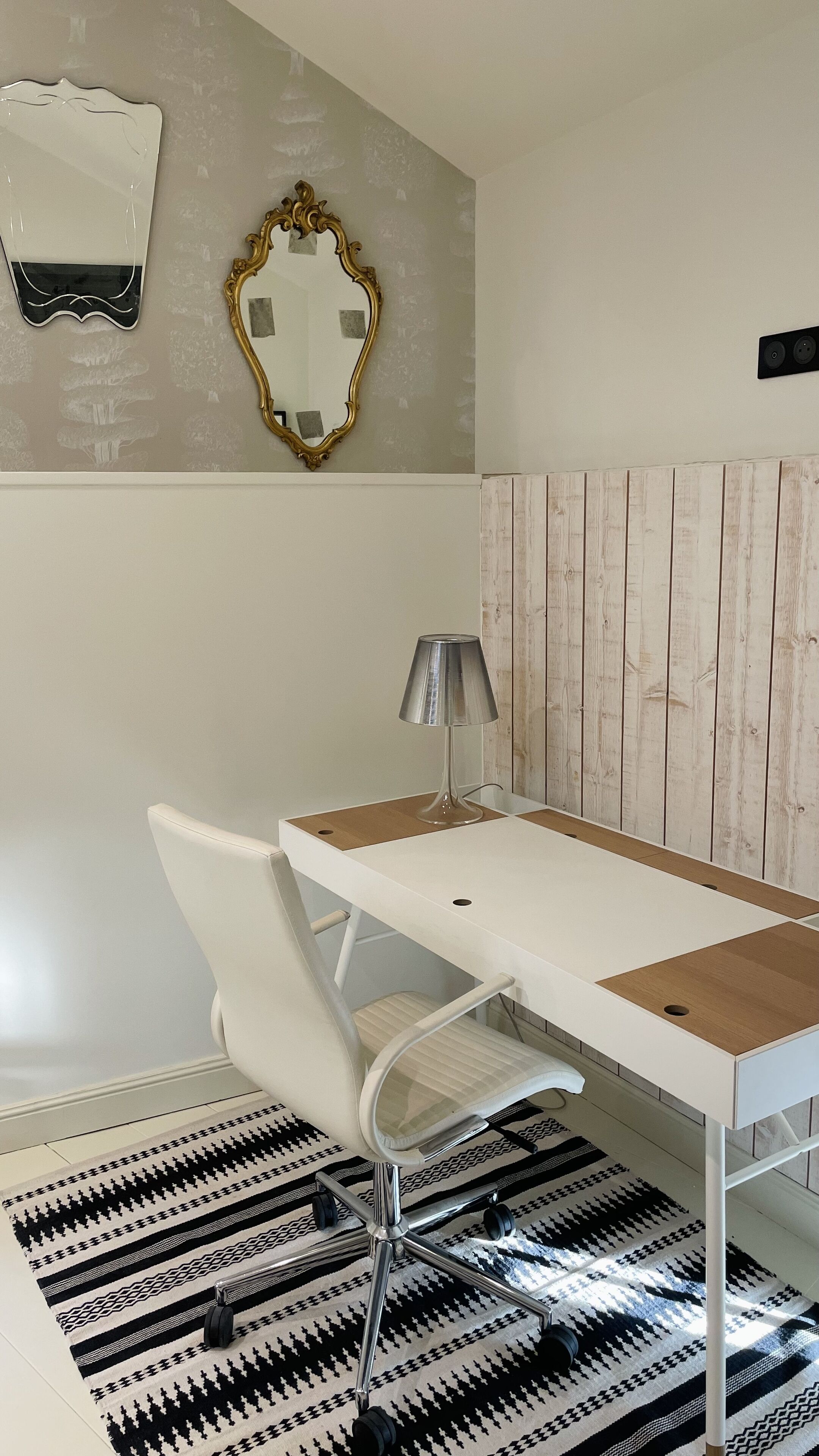 A modern room with a white desk, a white chair, a lamp, a patterned rug, and a wooden panel wall.