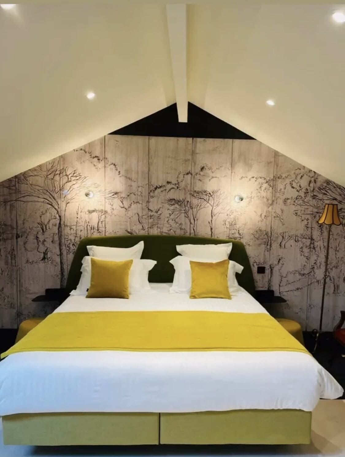 Un dormitorio con techo alto, cama con ropa de cama blanca y amarilla, y una pared con un mural de un árbol.