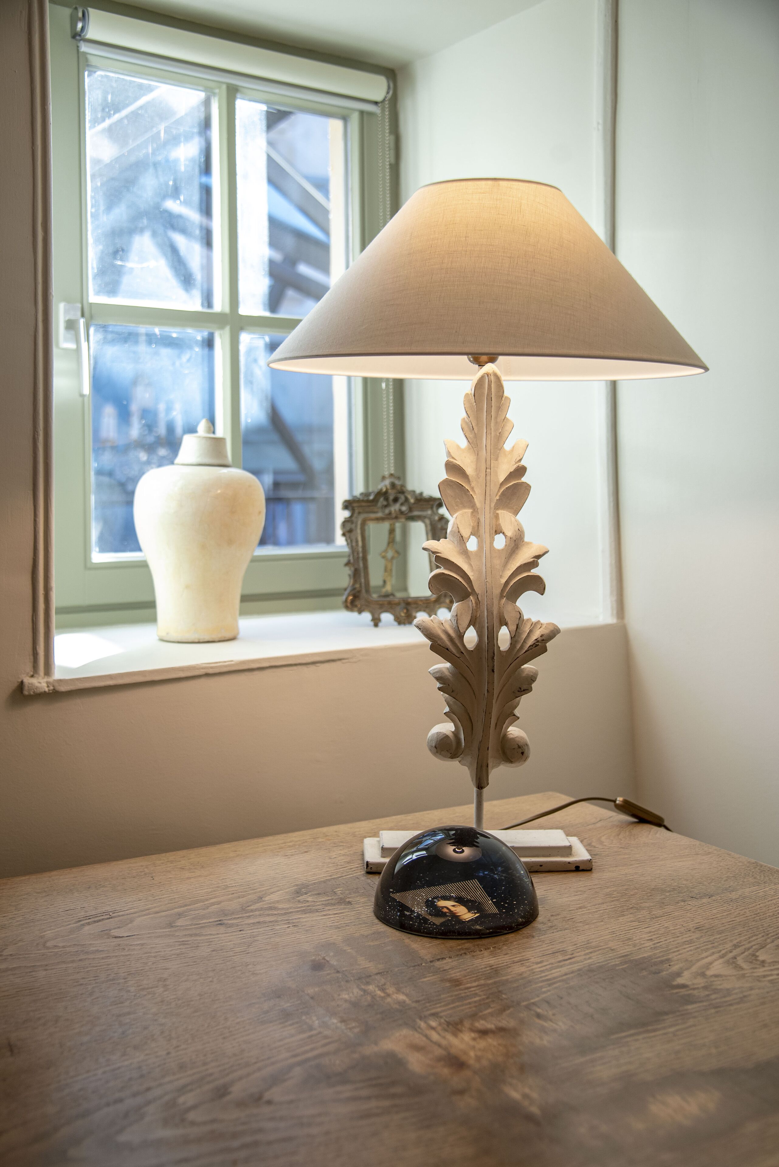 Une lampe de table avec un pied en forme de feuille et un abat-jour conique, placée sur une table en bois près d’une fenêtre. Sur le rebord de la fenêtre, on aperçoit un vase blanc et un objet décoratif.