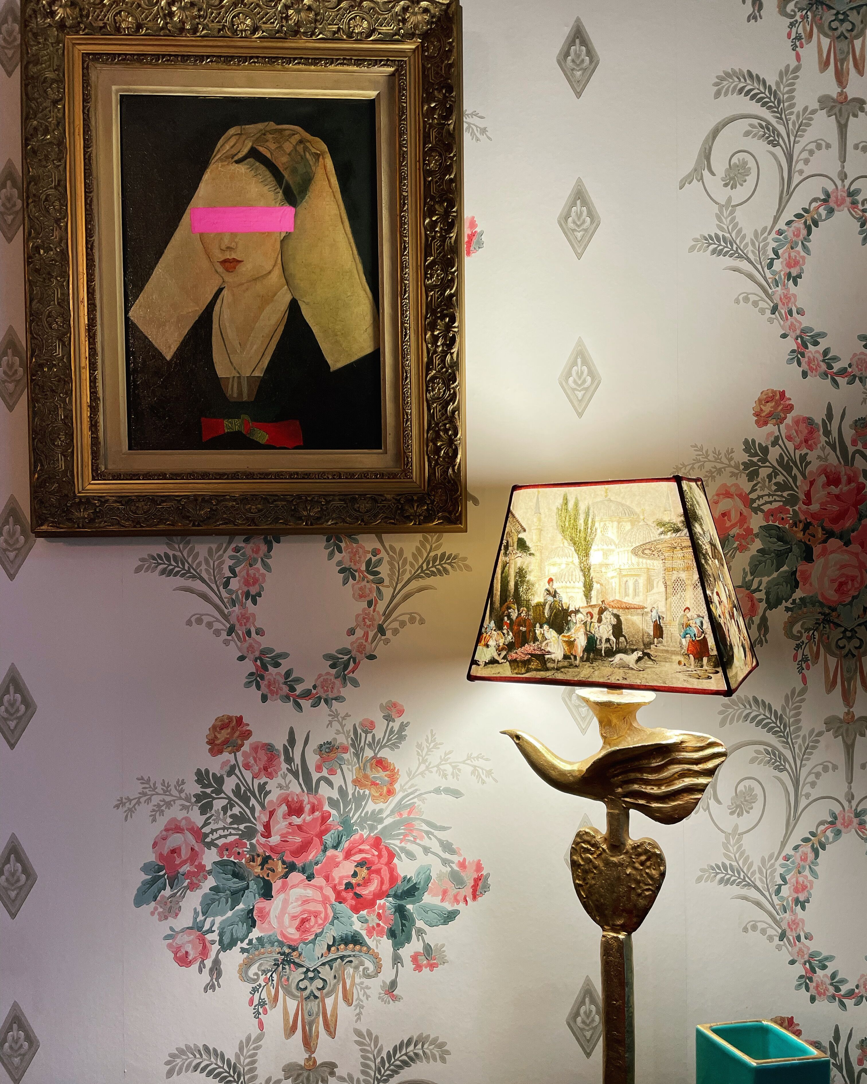 Un portrait encadré dont le visage est flou, une lampe avec un pied en forme d’oiseau et un papier peint à motifs floraux.