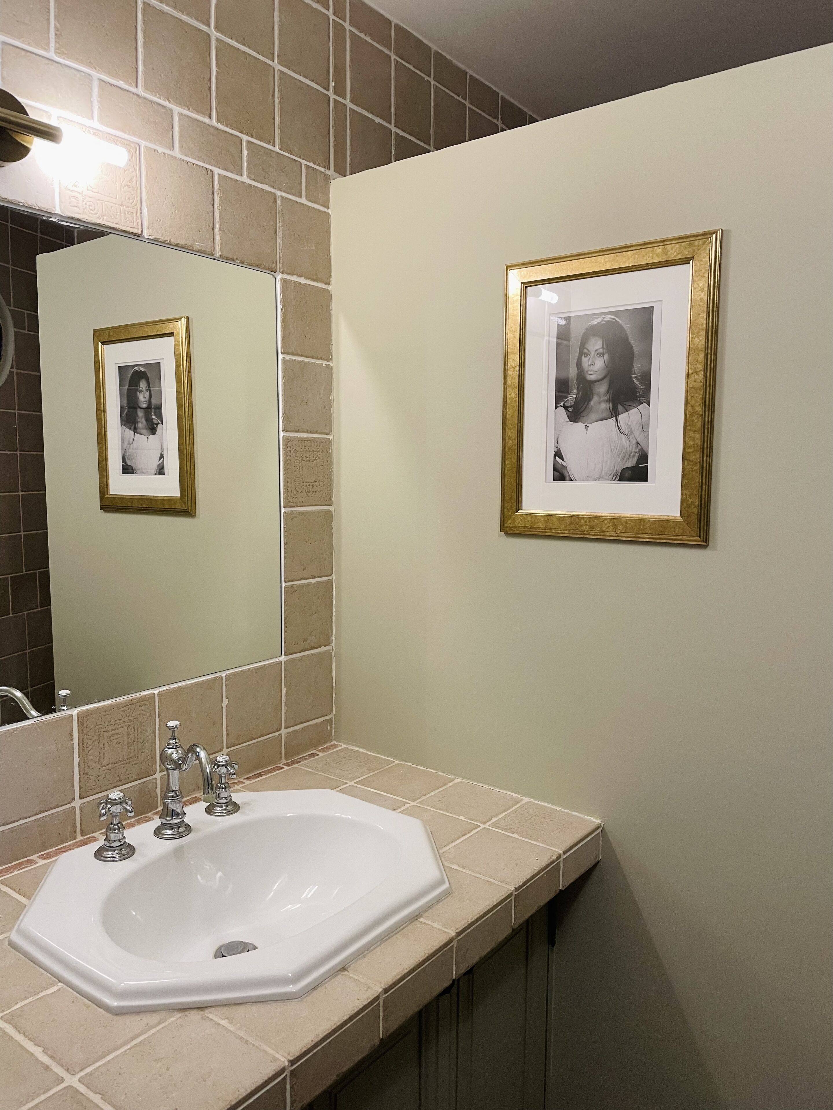 Un baño con un lavamanos blanco, dos grifos y un espejo. Hay dos fotos enmarcadas en la pared.