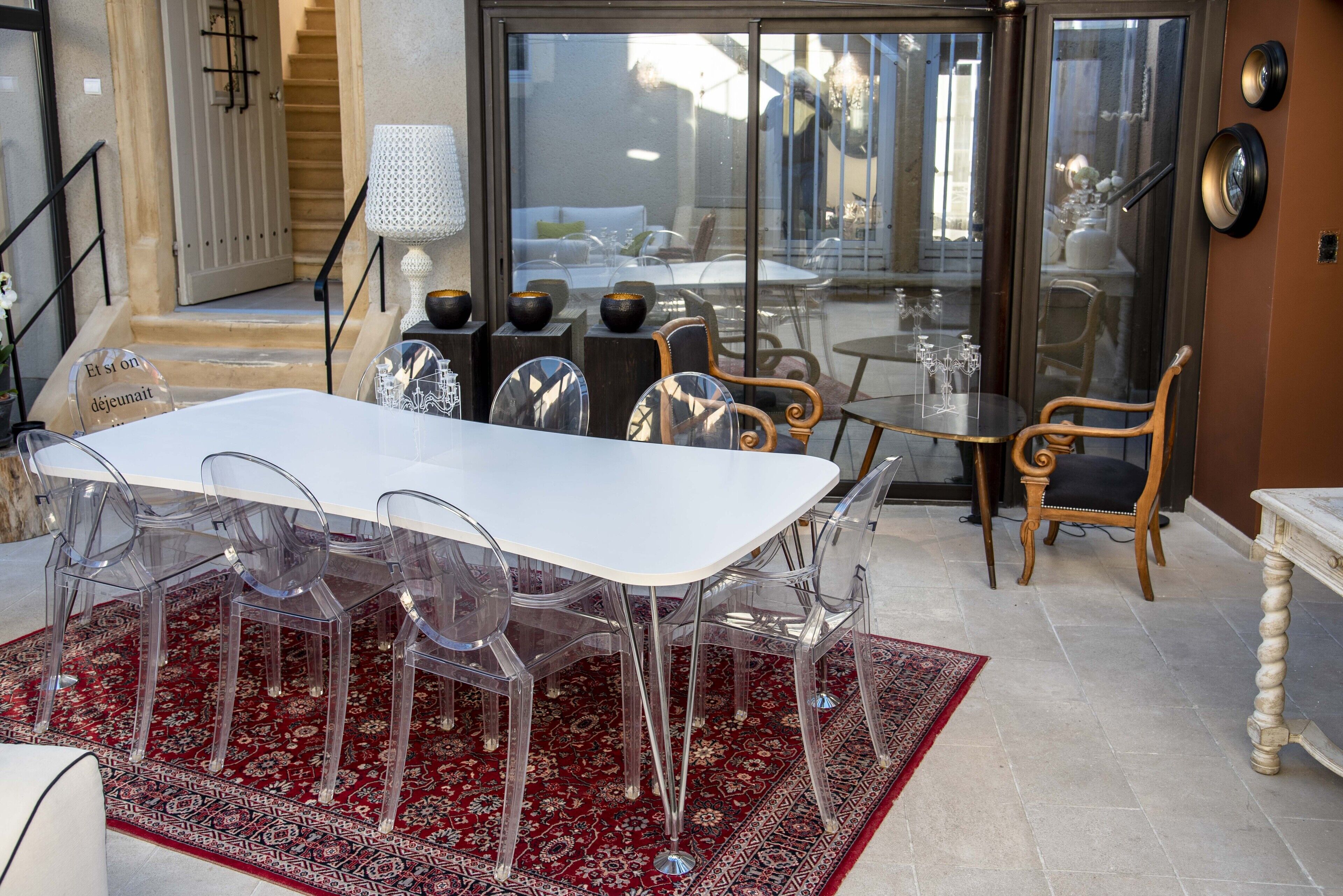 Un comedor con una mesa blanca y sillas transparentes, un tapete rojo estampado y vistas a través de puertas de cristal de un área de estar exterior.