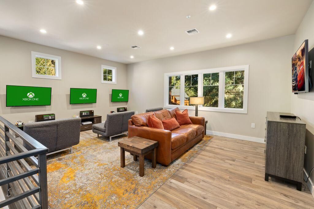 A modern living room with a brown leather sofa, grey armchairs, and a wooden coffee table. There are three green Xbox One displays on the wall.