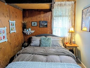 A cozy bedroom with a wooden wall, a bed with a knitted throw, a nightstand with a lamp, and framed pictures on the wall.