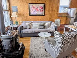 A cozy living room with a vintage stove, a grey sofa, patterned cushions, a houndstooth armchair, and a framed floral artwork on the wall.
