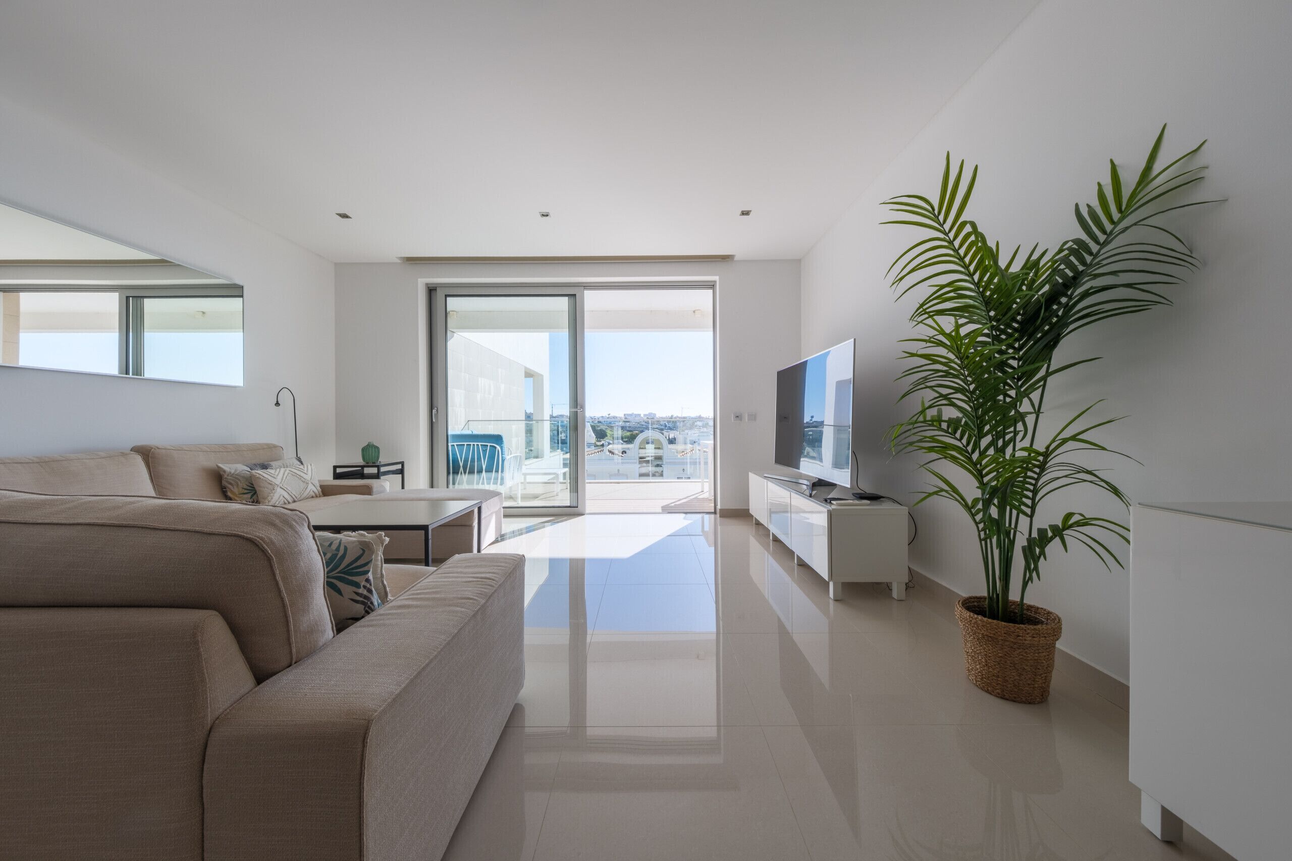 A modern living room with a sofa, a TV, a plant, and a balcony with a city view.