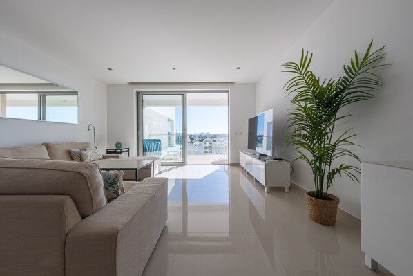 A modern living room with a sofa, a TV, a plant, and a balcony with a city view.