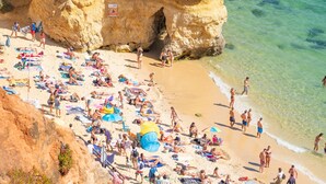 Een druk strand met mensen die zonnebaden en zwemmen, een grote rotsformatie en helder turquoise water.