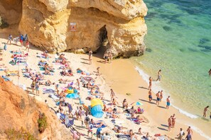 A crowded beach with people sunbathing and swimming, a large rock formation, and clear turquoise water.