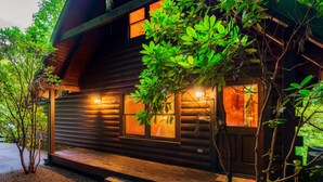 Une cabane en rondins avec une façade en bois, un porche et une fenêtre éclairée d’une lumière chaleureuse.