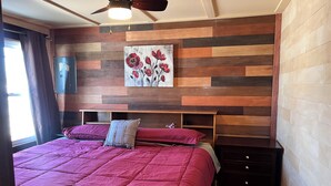 A bedroom with a wooden panel wall, a ceiling fan, a painting of flowers, a bed with a red bedspread, and a nightstand. - 4 Bedroom Apartment above Wilzke's Pub - Neighborhood Bar. (Menasha)