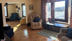 A living room with a beige armchair, a blue armchair, a wooden floor, a patterned rug, and a large window with curtains. - 4 Bedroom Apartment above Wilzke's Pub - Neighborhood Bar. (Menasha)