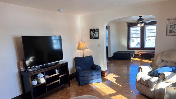 A living room with a flat-screen TV on a wooden stand, a blue armchair, a beige sofa, and a large window with natural light. - 4 Bedroom Apartment above Wilzke's Pub - Neighborhood Bar. (Menasha)