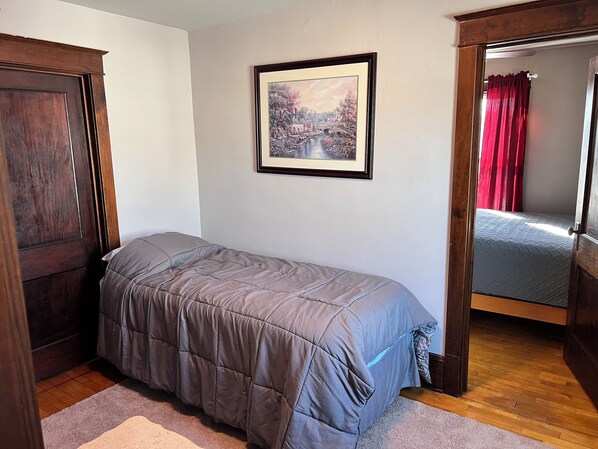 Un dormitorio con una cama individual, un armario de madera, un cuadro en la pared y vistas de una cortina roja a través de una puerta abierta.