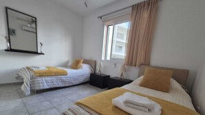Una habitación de hotel con dos camas, una ventana con cortinas, una mesita de noche con lámpara y un espejo adosado a la pared.