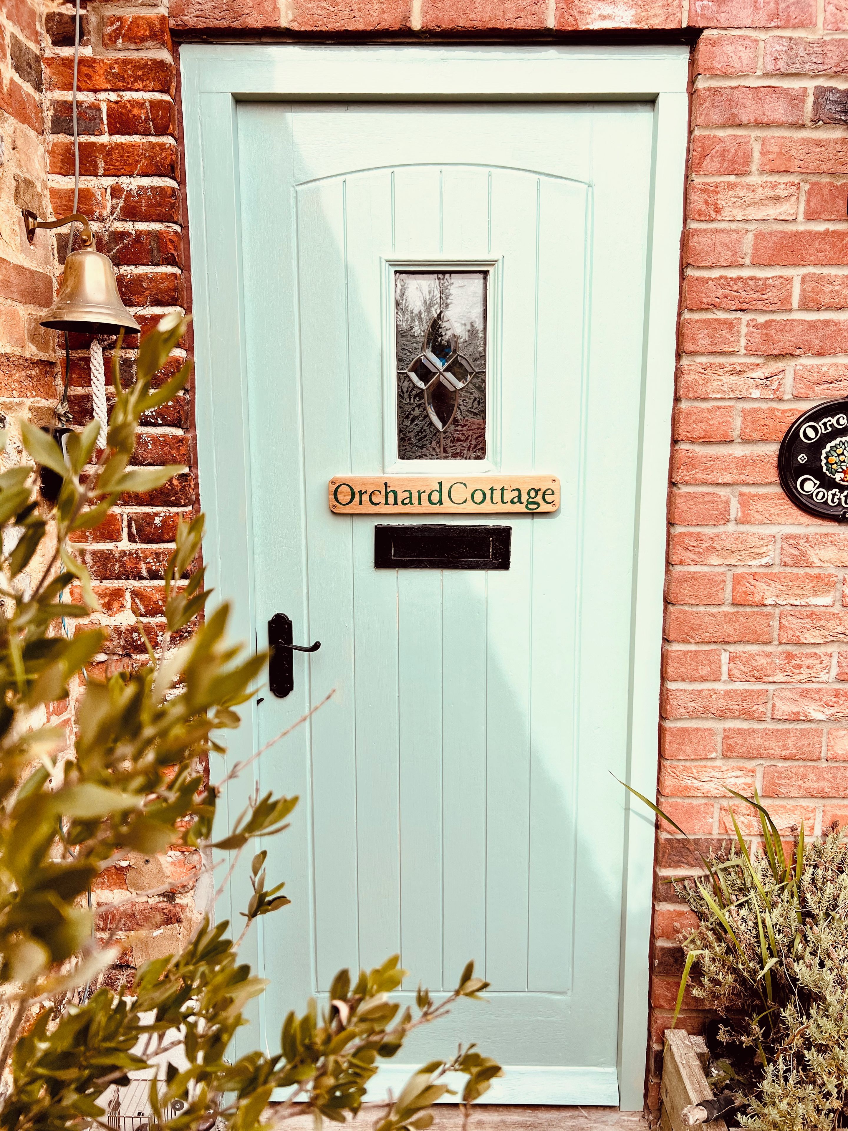 A light blue door with a small window and a bell above it. There is a sign above the door that reads 'Orchard Cottage.'