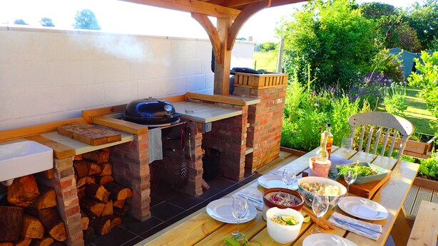 An outdoor patio with a brick barbecue, wooden tables, and a variety of dishes and drinks.