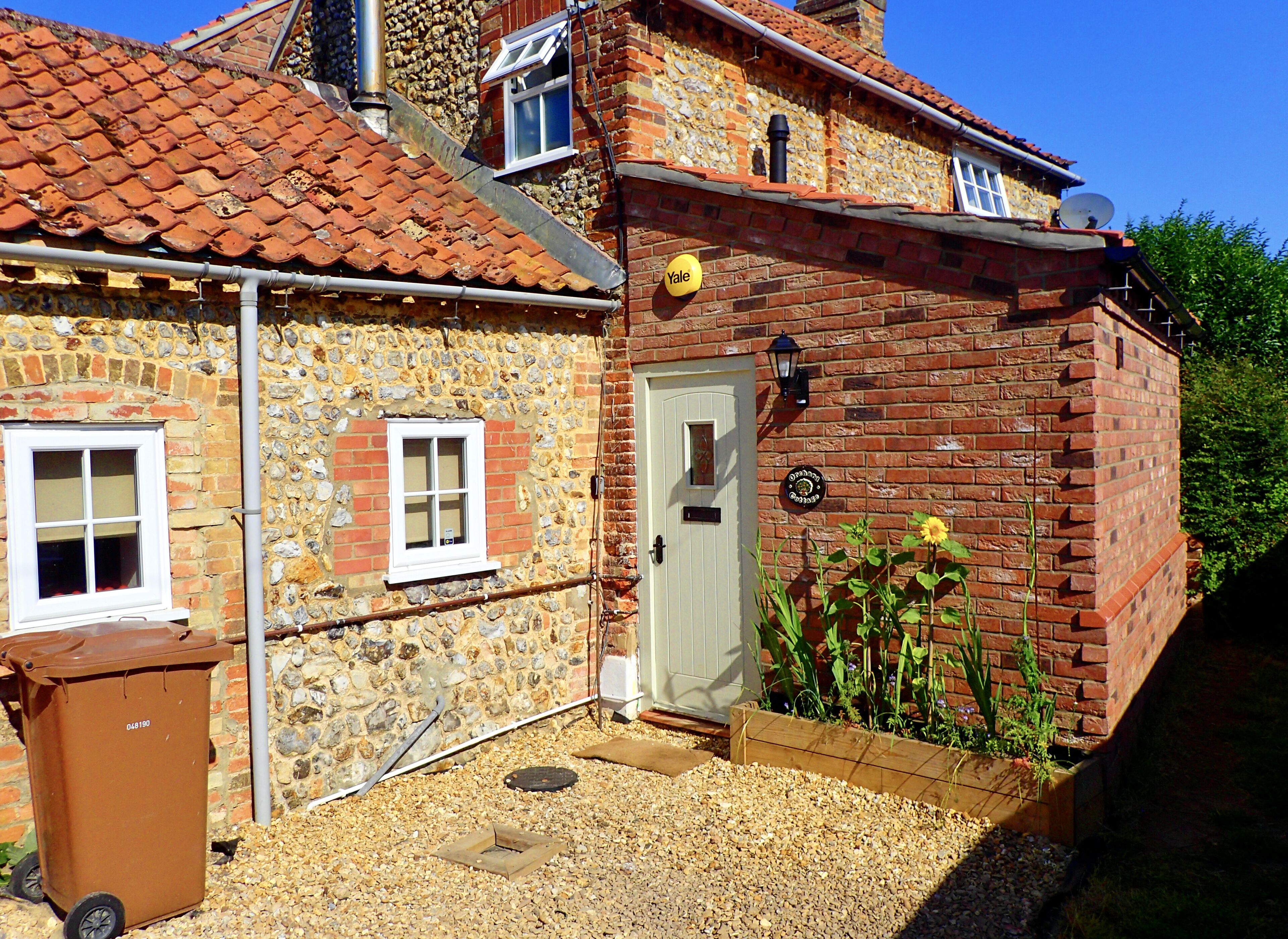 Une maison en pierre avec un toit recouvert de tuiles rouges, une porte blanche et un petit jardin planté de fleurs et d’arbustes.