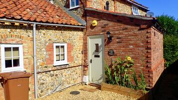 Une maison en pierre avec un toit recouvert de tuiles rouges, une porte blanche et un petit jardin planté de fleurs et d’arbustes.