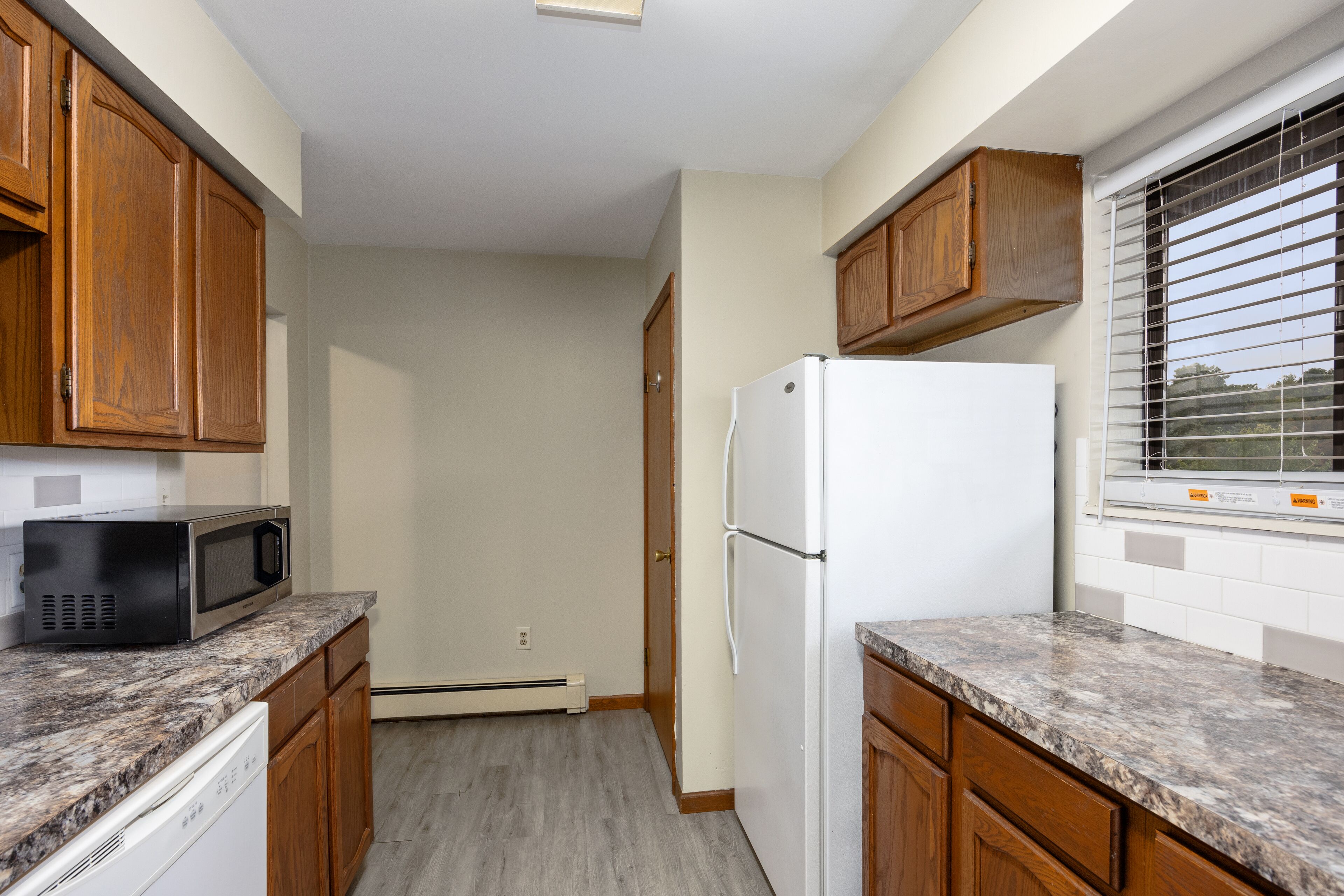 A kitchen with wooden cabinets, a white refrigerator, a microwave, and a window with blinds.