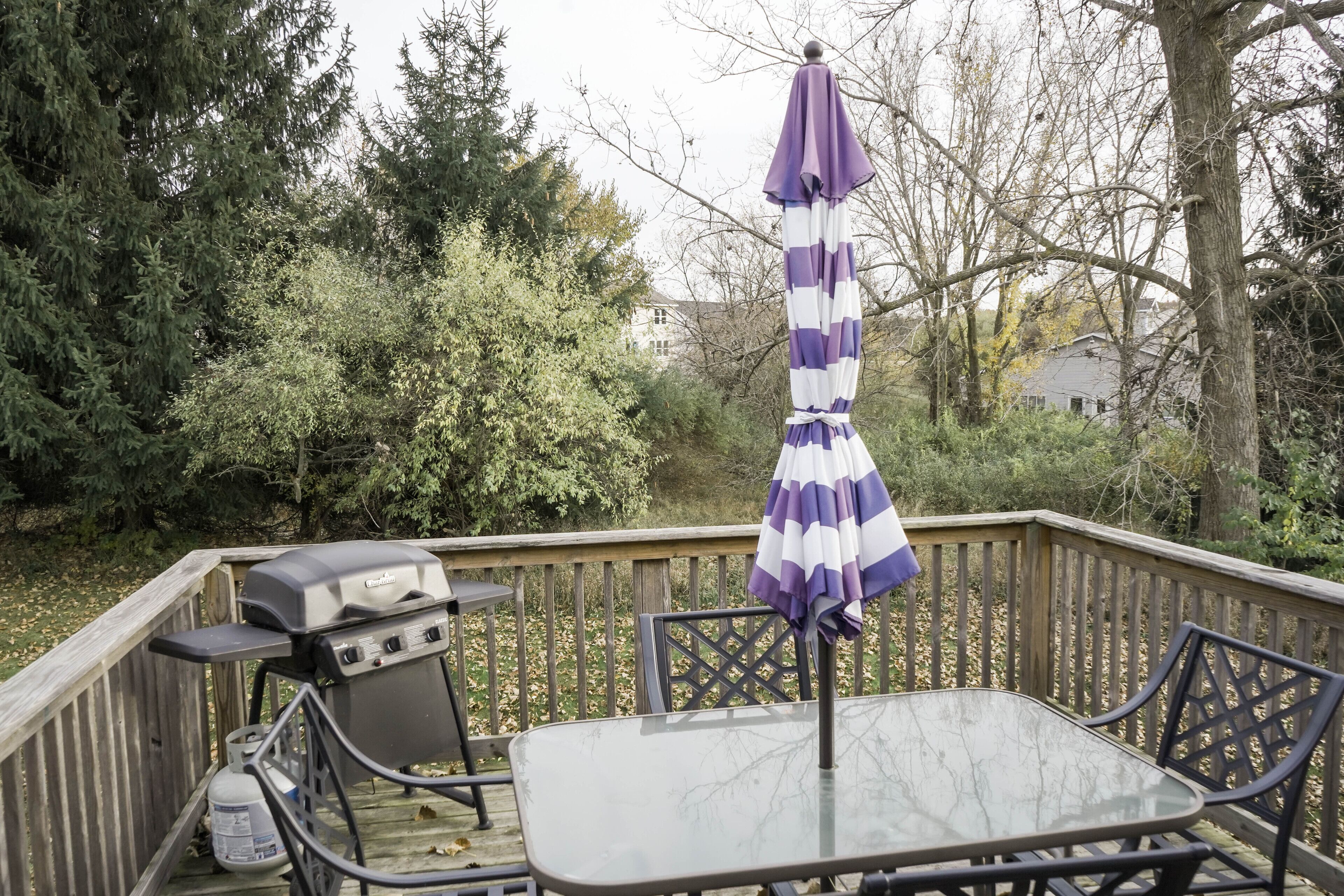 Una terraza con una parrilla, una mesa de vidrio y sillas, rodeada de árboles y una casa al fondo.