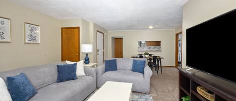 A living room with two grey sofas, a white coffee table, a flat-screen TV, and a kitchen area in the background.