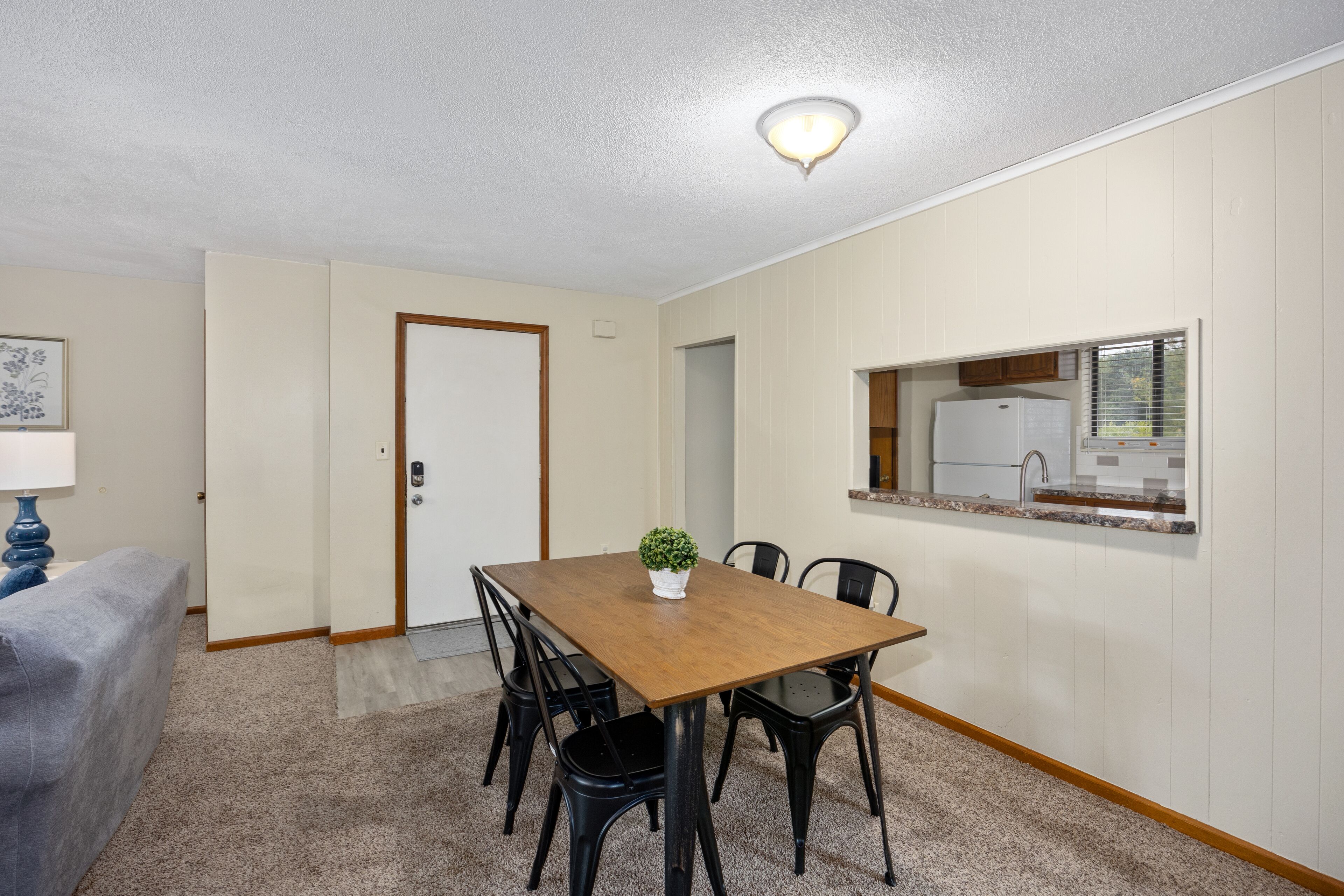 Un comedor con una mesa y sillas de madera, una planta pequeña en maceta y una cocina visible a través de una ventana.